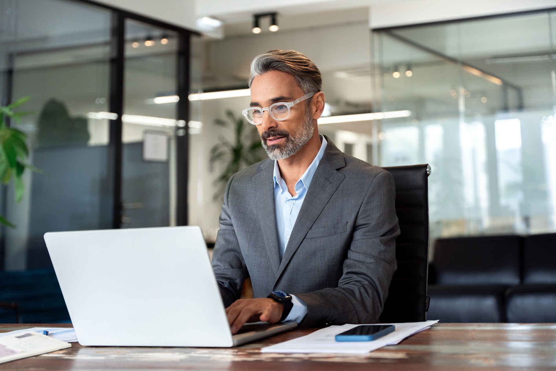 Mature Indian or Latin business man ceo trader using computer, typing, working in modern office, doing online data market analysis, thinking planning tech strategy looking at laptop with copy space.